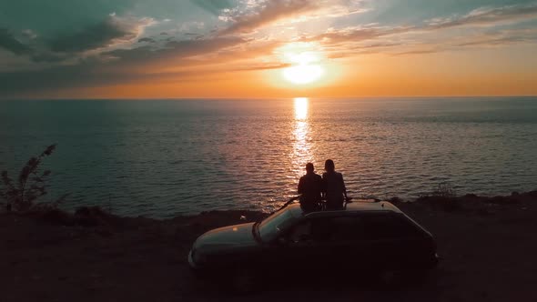 the couple sits on roof of car and watch sunset on sea