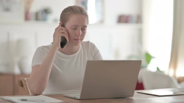 Beautiful Young Woman Talking on Phone While Using Laptop in Office