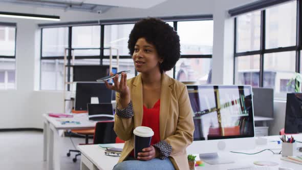 Mixed race businesswoman sitting on desk holding coffee talking on smartphone in office