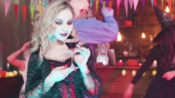 Young Beautiful Woman in Vampire Costume Drinks Blood From a Glass at a Halloween Party