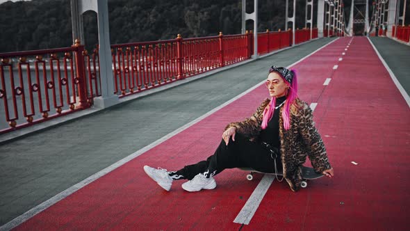Young Hipster Woman with Pink Hair Wearing Informal Clothes is Sitting Sideways on Skateboard Posing