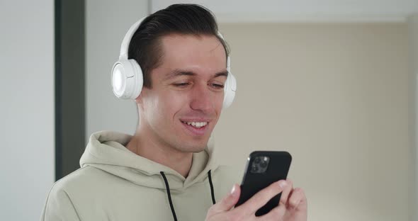 Closeup Portrait of Joyful Handsome Young Man Listening to Music in White Headphones and Surfing