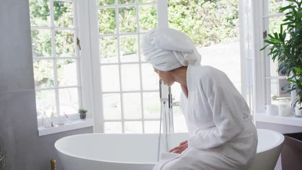 Mixed race woman checking water and sitting on bathtub in bathroom