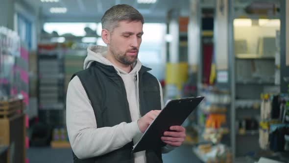 Concentrated Hardware Store Worker Standing with List of Products Checking Goods on Shelves