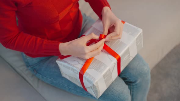 Close up hands of woman using ribbon to tie bow for a wrapped gift box at home.