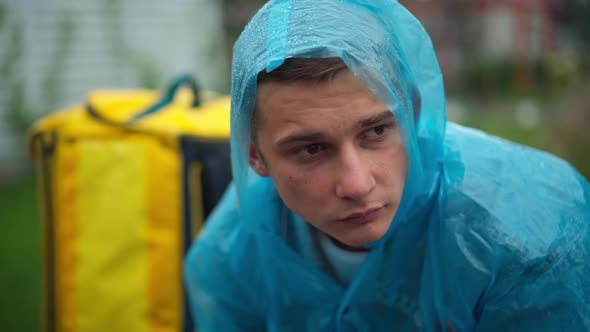 Closeup Portrait of Depressed Young Man in Rain Coat with Delivery Backpack Sitting Outdoors