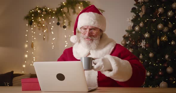 Cheerful Elderly Bearded Santa Claus Sitting at a Table Drinking Hot Drink While Working on Laptop