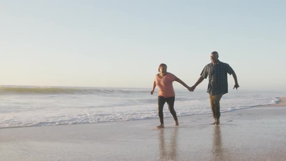 Smiling senior african american couple holding hands and running on sunny beach