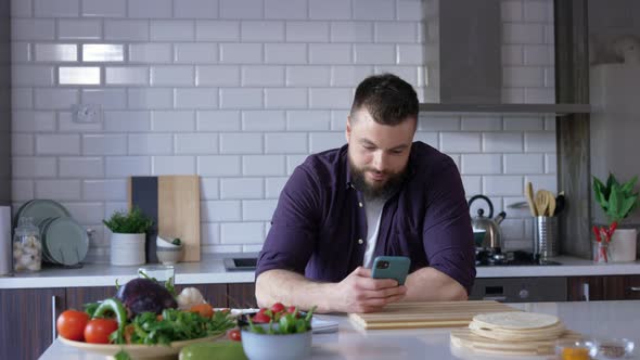 Handsome Young Man with a Smartphone in the Kitchen using Food App and Smiling