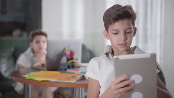 Close-up Portrait of Caucasian Boy Using Tablet As His Twin Brother Sitting at the Background