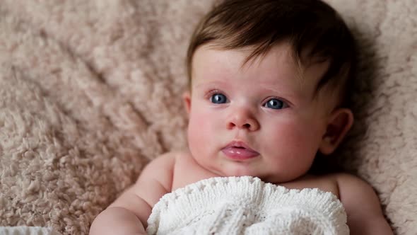 A baby girl is sitting on a wool covered pillow and smiling and putting her fist in her mouth