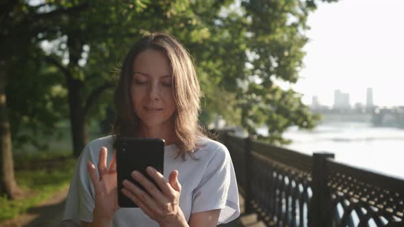 a Woman Walks and Looks Carefully at the Phone Screen on the City Embankment