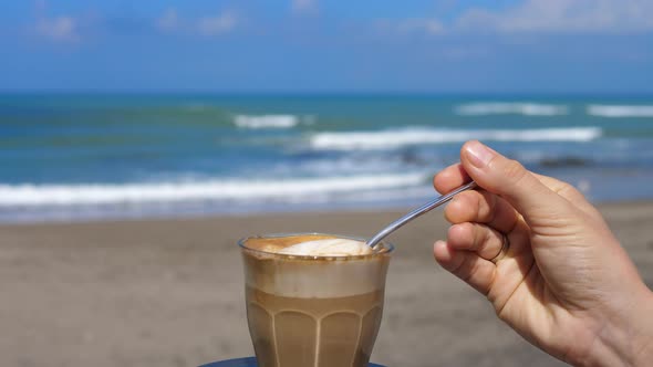 Joyful Coffee Break with Coffee with Milk Foam on the Seashore on the Vacation Hand Dipping Spoon in