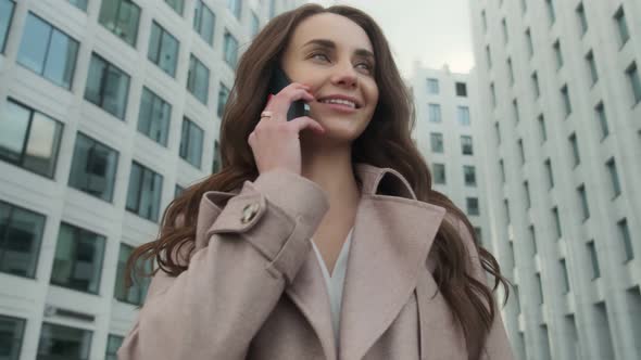 Cheerful young Caucasian woman standing in city street and texting on mobile phone with smile.