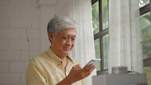 Senior Asian man talking on smartphone happy and smiling beside a window at home.