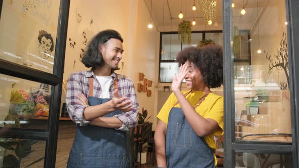 Two baristas at a cafe door, arms crossed, teasing together with a happy smile.