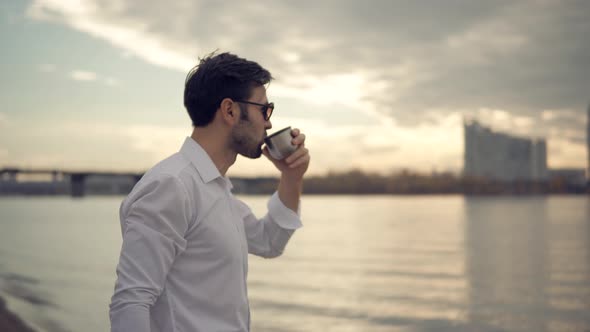 Man Relaxing On City Beach And Drinks Hot Chocolate. Man Drinking Tea From Thermo Cup. Cup Of Tea.