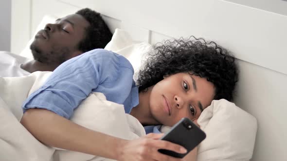 African Woman Using Smartphone While Boyfriend is Sleeping