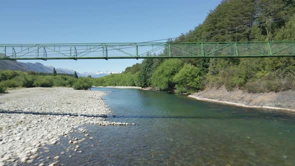 Aerial dolly in of Rio Azul stream flying under elevated bridge between trees and mountains, Patagon