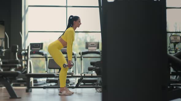 Fit Hispanic Woman Performing Weight Lifting Deadlift Exercise with Dumbbell at Gym In Yellow