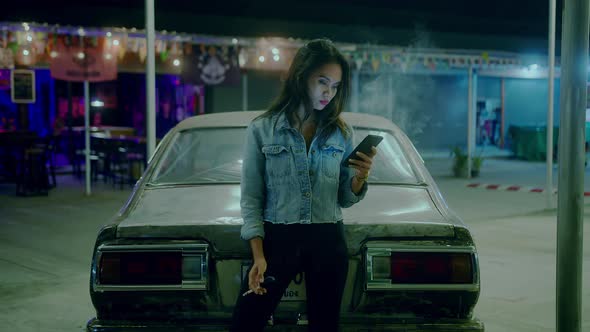 A Girl Smokes in Front of a Rusty Classic Car While Typing a Message Late at Night