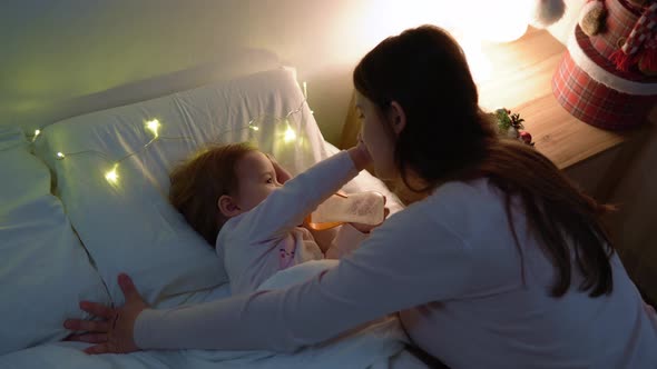 Young Mother Feeding Nutrition Breast Milk to Little Baby Girl From Bottle Lying on Bed in Bedroomat