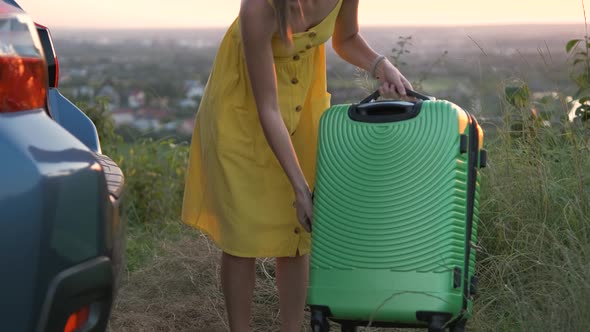 Young woman in yellow dress putting green suitcase in car trunk. Travel and vacations concept.