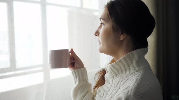 Young Beautiful Brunette Woman in Sweater Drinking Cup of Coffee Near the Window