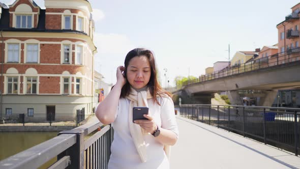 Asian black straight hair woman walking on a bridge over a river in town and using her smartphone