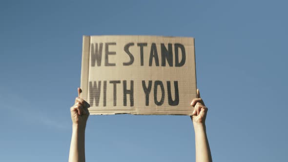 Sign WE STAND WITH YOU against blue sky. Woman hands waving poster "We Stand With You".
