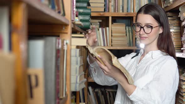 Young Attractive Student in Glasses Turns Pages in the Book Which She Took From Books on Shelves in
