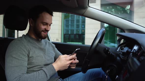 Happy smiling young man sitting in car and typing online message on cell phone, side view.