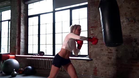 a Blonde with Long Hair in a White Top and Red Boxing Gloves Hits a Punching Bag Against the