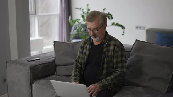 Senior Older Mature Man Typing on Laptop Browsing Internet Sit on Sofa Alone