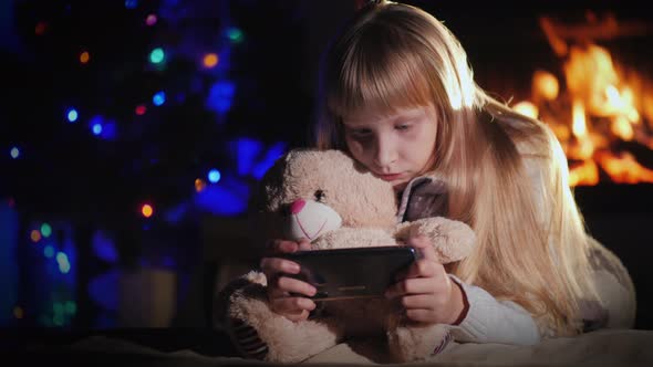 A Child with a Teddy Bear Is Playing on a Smartphone Against the Background of a Fireplace