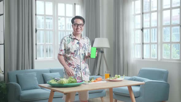 Asian Man With Healthy Food Holding And Pointing Green Screen Smartphone At Home