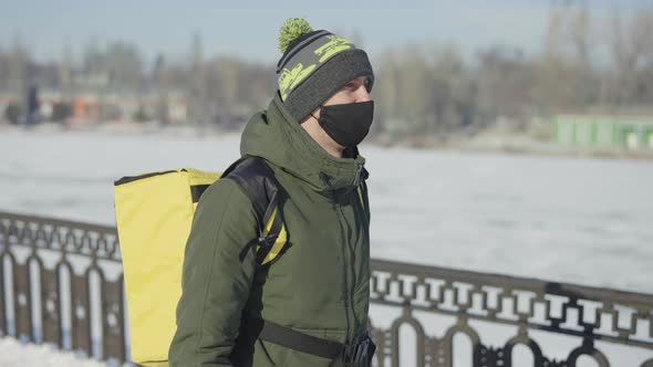 Courier in Medical Mask with Yellow Thermo Bag Walking Along Snow Covered City Street