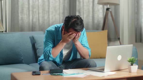 Asian Man With A Laptop Looking At The Bill And Having Headache Due To Financial Problem