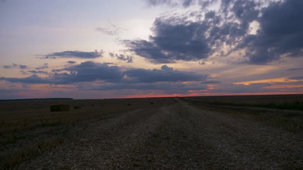 Timelapse of cloudy orange sunset sky over harvest field with haystack