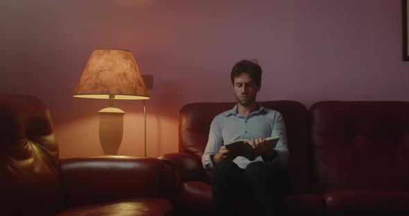 Young guy with shirt reads a book sitting in the living room at home