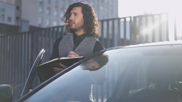 Young Focused Man Writing with Pen Standing in Sunlight at Black Car in Urban City