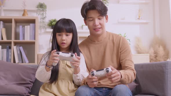 Happy Asian father teaching daughter to play games with joy stick on sofa in relax day.