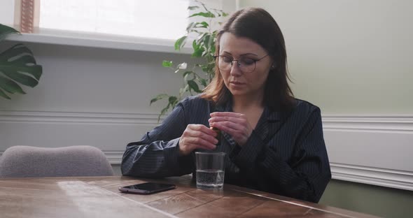 Mature Woman Dripping Drops with Glass of Water, Drinking Sedative Medicine, at Home in Pajamas