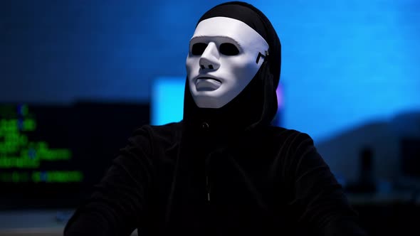 Middle Shot of Man in Black Hoodie and Anonymous White Mask Looking Around in Blue Room and Typing