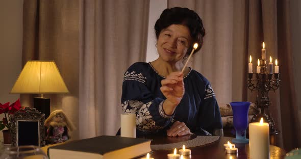 Front View of Smiling Caucasian Brunette Woman Sitting at the Table and Lighting Up Candles. Senior