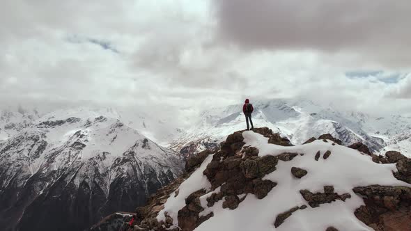 A Male Traveller with a Backpack Stands on Top of a Cliff Against the Backdrop of Snow-capped