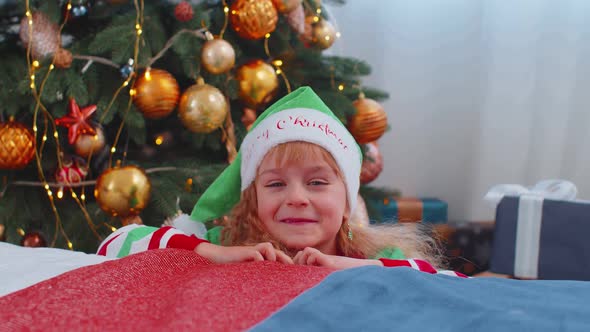 Toddler Girl in Santa Claus Elf Pajama Hiding Behind Bed Playing Hide and Seek Game Christmas Tree