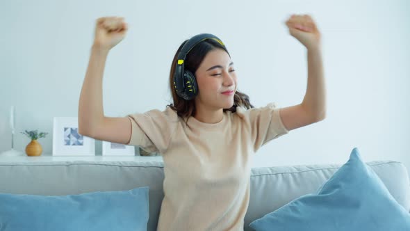 Asian beautiful woman put on headphone feeling happy listen to music and dance on sofa at home.