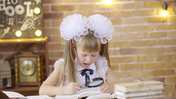 Little Girl Reads a Book with Magnifier Sitting at Table on Background of a School Class