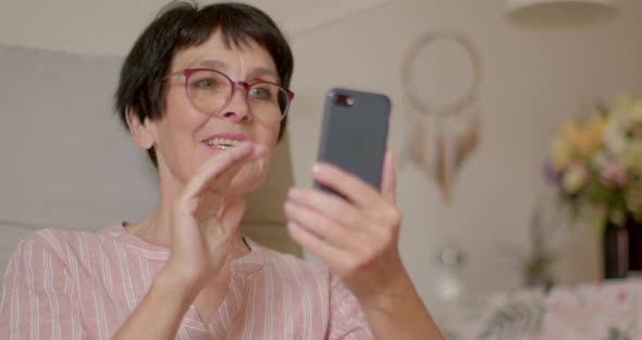 Happy Aged Woman Uses Phone Smiles Laughs Waves Her Hand and Sends Air Kiss